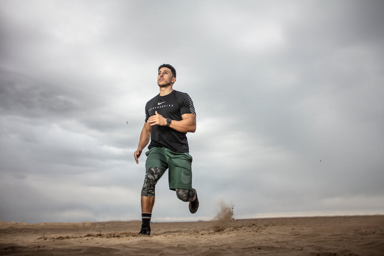 hero-img-02 A muscular man running on sandy terrain under cloudy skies, promoting fitness and adventure.